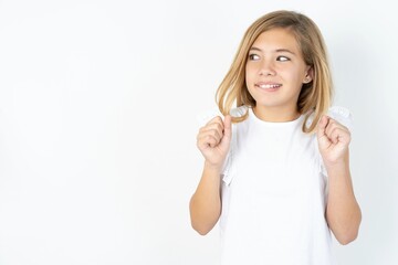 beautiful caucasian teen girl wearing white T-shirt over white wall clenches fists and awaits for...