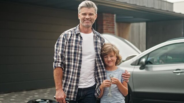 Man and his son standing in yard. Happy family members are looking at camera, posing and smiling outdoor. Young cute boy holding wrench and enjoy spending time with dad. Father hugs his child.