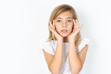 Happy beautiful caucasian teen girl wearing white T-shirt over white wall touches both cheeks gently, has tender smile, shows white teeth, gazes positively straightly at camera,