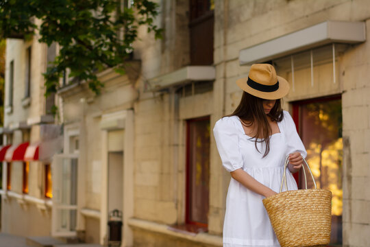 Elegant young woman looking in her wicker bag for a phone in a European city. Stylish woman a white dress, a straw hat, a wicker basket on the street. Street style summer, fashionable outfit