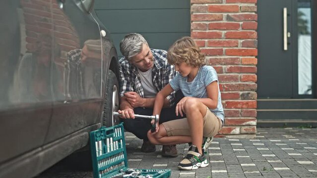 Portrait of pre-adolescent good boy helps dad repair car. Father and his little son spending time together outdoor in yard. Adult male guardian brings up and raiss child. Concept of happy childhood.