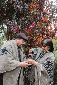 Serious mature couple in traditional Mapuche clothes with thermos and mug pouring hot yerba mate while standing in autumn garden in Temuco
