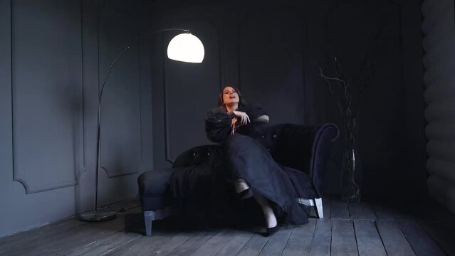 Woman with dark hair wearing lovely black dress emotionaly sings on black sofa