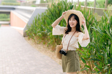 Portrait of asian young woman traveler with weaving hat and basket and a camera on green public park nature background. Journey trip lifestyle, world travel explorer or Asia summer tourism concept.