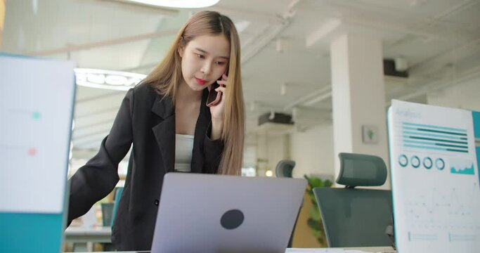Young busy asian business woman working on project paperwork talking phone, using laptop remotely work in modern office. Female freelancer entrepreneur making distance job telesale call with customer.