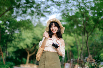 Portrait of asian young woman traveler with weaving hat and basket and a camera on green public...