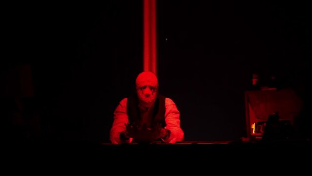 A man in a mask of an evil being sits at a table. In the hands of another mask. Twilight. Red light. Sad evil clown mask without mouth. High quality FullHD footage
