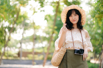 Portrait of asian young woman traveler with weaving hat and basket and a camera on green public park nature background. Journey trip lifestyle, world travel explorer or Asia summer tourism concept.