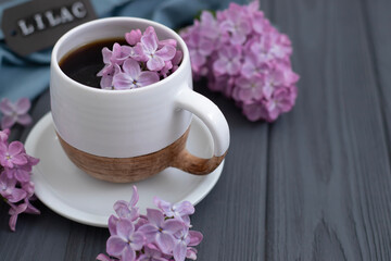 A cup of Americano black coffee, purple lilac flowers on a wooden background. A cup of Americano black coffee, purple lilac flowers on a wooden background. Lilac inscription on the card next to it