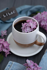 A cup of Americano black coffee, purple lilac flowers on a wooden background. A cup of Americano black coffee, purple lilac flowers on a wooden background. Lilac inscription on the card next to it