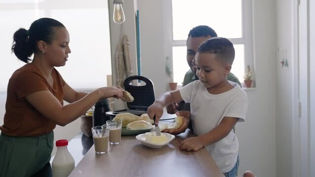 Cheerful mixed race family making breakfast while husband and little son eating and drinking juice at kitchen table
