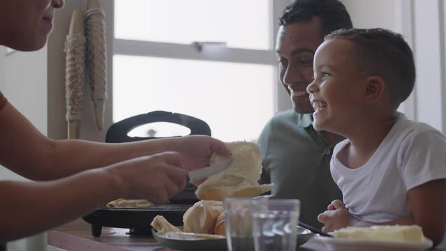 Cheerful mixed race family making breakfast while husband and little son eating and drinking juice at kitchen table
