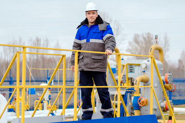 Portrait of worker in white helmet and winter pea jacket at industrial gas facility. Middle-aged Caucasian man stands in full growth on territory of gas distribution station. Genuine worker.