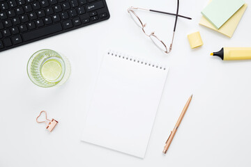 Workplace with white notebook, black keyboard, stationery, glass of water with lime and glasses on white desk. Flat lay office desk, mock up space for text. Top view. Copy space