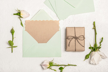 Feminine invitation or greeting card mockup with light green craft envelope, gift box and white roses on light background.
