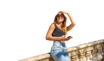 Beautiful tanned young woman in a black top and jeans holding her mobile phone and covering her eyes from the sun looking for someone resting on a fence of granite in PNG background