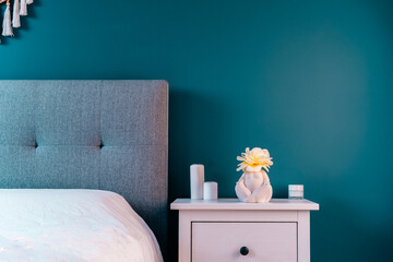 Cozy bedroom with turquoise blue walls, white bedside table with ceramic female body-shaped vase with flower and candles. Dark modern stylish female room. Self recovery place at home. Selective focus.