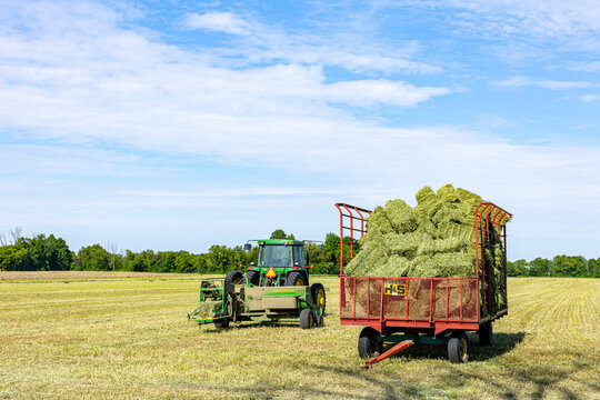 A John Deere tractor and small square baler next to an H&S hay wagon full of bales in a big field.