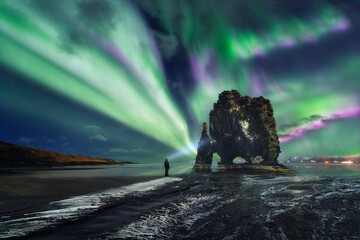 Aurora borealis above Hvitserkur rock formation, A man with a lantern (headlamp) is watching dance...