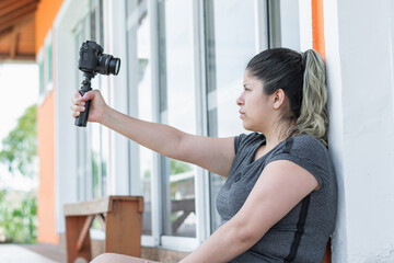 young latina woman blogger, sitting on the ground next to the house, with camera in hand recording herself
