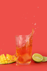 Glass of tasty cider with splashes, ice cube, mango and lime on red background