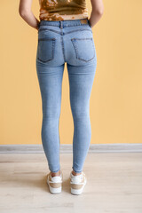 Young woman in stylish jeans near yellow wall, back view