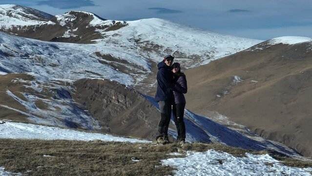couple standing on top overlooking the snow-capped alpine mountains. A man and a woman stand hugging on a field among high mountains in the snow, on a sunny day, flying around people on a drone