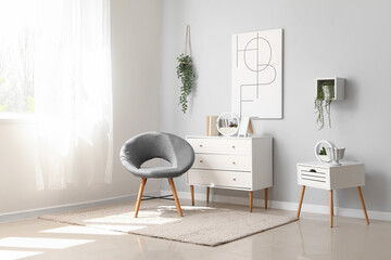 Interior of light living room with cozy grey armchair, chest of drawers and table