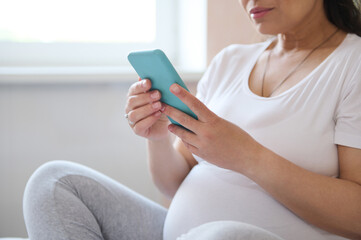 Close-up beautiful adult pregnant woman using smartphone, checking mobile app, sharing maternity lifestyle on social media, sitting on the bed in a cozy apartment. Happy carefree pegnancy. Copy space