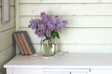 Table in a wooden cottage concept. A bouquet of purple lilac in a glass jar, old books.