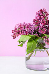 Glass vase with beautiful lilac flowers on table against pink background