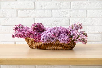 Schilderijen op glas Wicker basket with beautiful lilac flowers on wooden table near light brick wall, closeup © Pixel-Shot