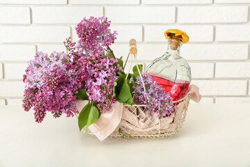 Basket with bottle of liquor and lilacs on white table near brick wall