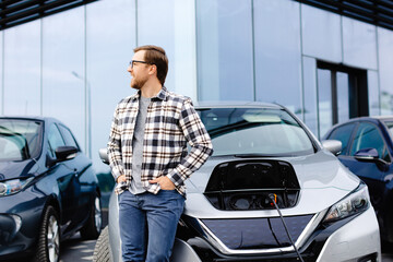 A young man stands next to his new electric car and rests while the car is charging