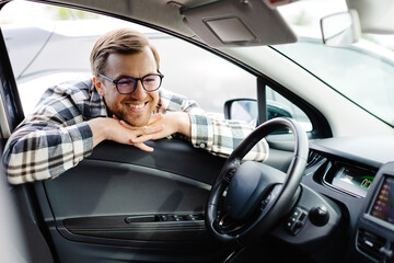 Young man looking through window inside new car, checking it before purchase at modern dealership store. Cheerful guy buying or renting automobile at showroom