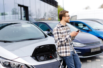 A young man uses a mobile phone while leaning on his electric car while the car is charging