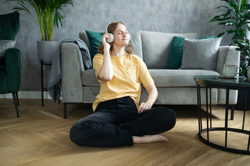 Obraz premium Positive young lady in wireless headphones and casual clothes listening to music with pleasure sits near a sofa in light modern living room. High quality photo