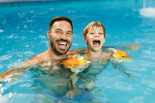 Playful father having fun with his little boy while being in swimming pool. There are looking at camera