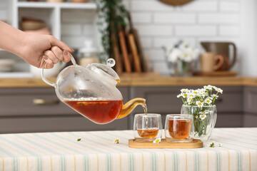 Female hand pouring natural chamomile tea from teapot into cup on table in kitchen
