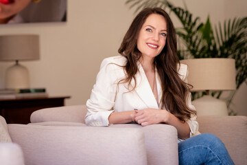 Obraz premium Portrait of brunette haired young woman wearing white blazer and relaxing in an armchair at home