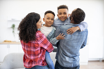 Fototapeta premium Back view of multicultural spouses holding preschool boys in arms while having break from playing board games. Friendly family planning after-dinner walk before leaving cozy home on sunny day.
