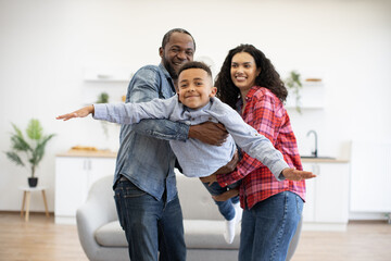 Fototapeta na wymiar Active african american adults holding happy boy in arms together while testing imaginary flight at home. Energetic kid in jeans wear experiencing feeling of freedom and well-being thanks to parents.