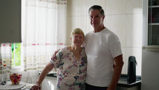 Middle-aged adult son posing for camera with his elderly mother in 80s standing in kitchen at home. Authentic domestic lifestyle of real life intergenerational people