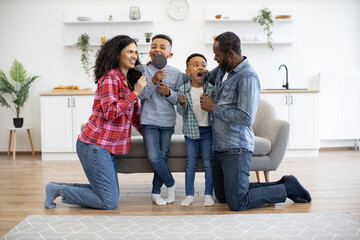 Joyful multicultural woman and man standing on knees near sofa with boys on it while pretending to take part in karaoke. Artistic parents and playful sons singing with kitchen utensils as microphones.