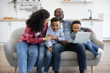 Affectionate african american mom tickling one of cute sons while happy father showing video to another boy. Cheerful parents and children using wireless laptop while having fun at home.