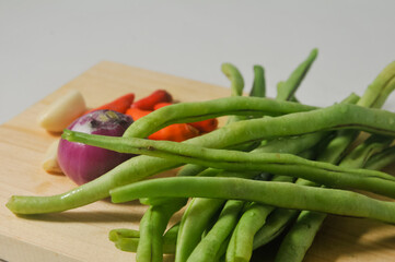 Some green beans, some red chili peppers, two cloves of garlic and a red onion on a wooden chopping board isolated on a white background