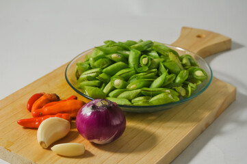 Sliced green beans on a small plate, some red chili peppers, a few cloves of garlic, and an onion on a wooden chopping board isolated on a white background