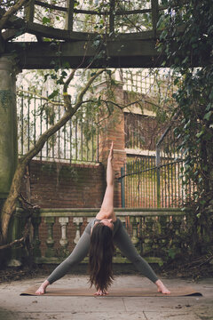 Vertical photo of a woman yoga teacher in Revolved or Twist Wide Legged Forward Bend Pose in a terrace of a historic landscaped garden with vegetation and stone classic columns. Outside.