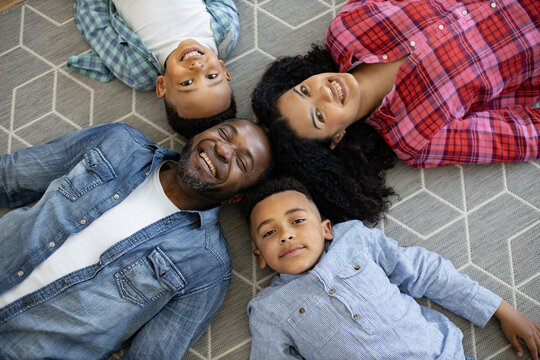Top view of multiethnic four-person family lying in circle with heads together on floor. Smiling parents and joyful sons sharing enormous positive energy in home interior.