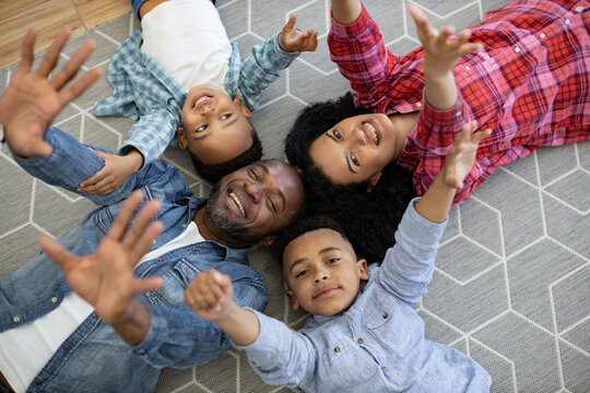 Top view of multicultural spouses and children making circle with their heads on gray rug with hands raised up. Smiling family of four expressing joy and excitement while spending time together.